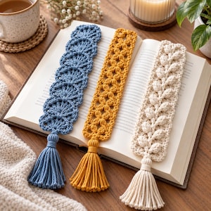 May include: Three rectangular crochet bookmarks resting on an open book. The left bookmark is blue with a scalloped pattern, the center is mustard yellow with a floral motif, and the right is cream with a leaf design. Each has a matching tassel.