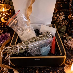 Peut inclure: Une boîte cadeau noire avec des bordures dorées contenant un bâton de sauge, un sachet en plastique de poudre blanche, une petite bougie rouge, une enveloppe blanche et un sachet en plastique avec une pierre bleue lisse, sur du papier noir.