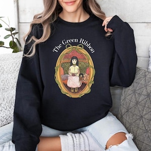 Op de afbeelding: Een zwart sweatshirt met lange mouwen en een grafische print van een persoon in een geel shirt en roze rok zittend in een stoel, omlijst door een groen lint en een ovale rand, met de tekst The Green Ribbon boven de illustratie.