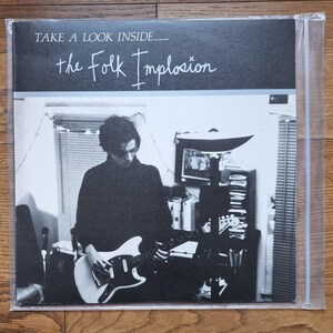 Pode incluir: Uma capa de disco de vinil intitulada Take A Look Inside de The Folk Implosion. A capa em preto e branco mostra uma pessoa tocando uma guitarra elétrica em um quarto com uma luminária, prateleiras e persianas. Texto branco em uma faixa preta.