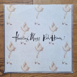 Pode incluir: Uma capa de disco de vinil quadrada com um padrão repetitivo de pernas ilustradas em castanho claro sobre um fundo branco sujo texturizado. O texto Throwing Muses Red Heaven está impresso ao centro numa caligrafia preta estilizada.
