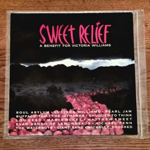 Pode incluir: Uma capa de álbum de música quadrada intitulada Sweet Relief: A Benefit for Victoria Williams. A arte apresenta uma paisagem escura com um caminho sinuoso de morangos vermelhos em primeiro plano e uma lista de artistas em branco.