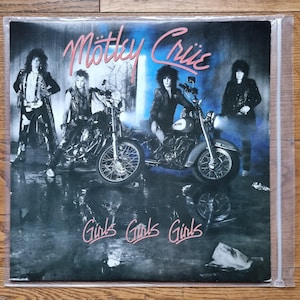 Può includere: Una copertina di un disco in vinile con la band Mötley Crüe. L'immagine mostra quattro membri della band, due in piedi e due seduti su motociclette, su uno sfondo scuro e suggestivo. Il testo Mötley Crüe appare in rosa in alto, e Girls Girls Girls è stampato in rosa in basso.