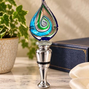 May include: A bottle stopper featuring a teardrop-shaped glass topper with swirling patterns of blue, green, and silver metallic tones. The base is made of polished chrome-coloured metal with a black rubber ring for a secure fit in a bottle neck.