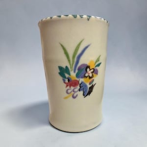 May include: A cylindrical ceramic tumbler with an off-white base colour. The exterior features a hand-painted floral motif in shades of green, blue, yellow, purple, and red. The top rim has a scalloped edge with small, evenly spaced dark markings.