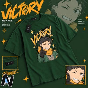 May include: A dark green short-sleeve t-shirt featuring a graphic of an anime character with dark hair, a yellow scarf, and black gloves. The text VICTORY is printed in yellow above the character. A black tag with a plus sign is attached to the collar.