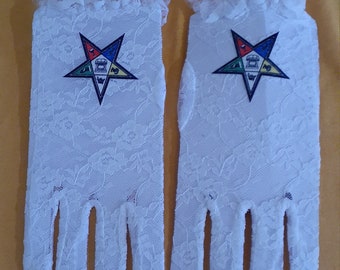 May include: A pair of white lace gloves with a Masonic symbol embroidered on each glove. The symbol is a five-pointed star with a compass and square in the center.