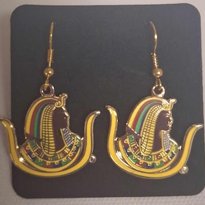 May include: Gold tone earrings with a colorful image of an Egyptian queen wearing a crown and a yellow and gold headdress. The earrings are dangling from gold hooks.