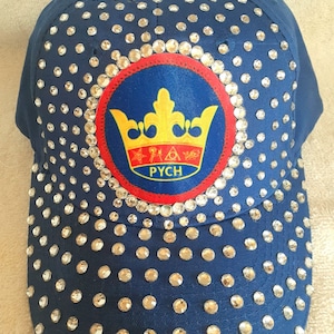 May include: A navy blue baseball cap with a large, round, red, yellow, and blue patch featuring a gold crown and the word "PYCH". The cap is covered in clear rhinestones.