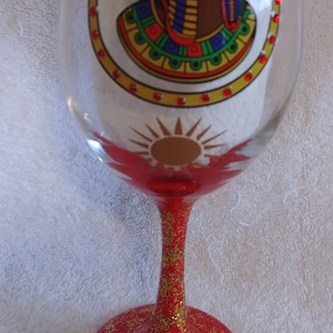 May include: A red wine glass with gold glitter on the stem and base. The glass features a painted design of an Egyptian queen with a sun behind her.