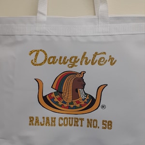 May include: White tote bag with gold glitter text that reads "Daughter" and a gold image of an Egyptian queen. The text "RAJAH COURT NO. 58" is also in gold glitter.