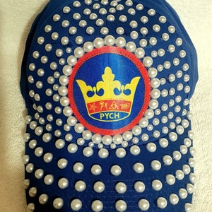 May include: A blue baseball cap with a crown and the word "PYCH" embroidered on a patch. The cap is covered in white pearl-like embellishments.