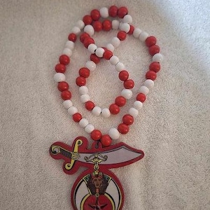 May include: A necklace with red and white wooden beads. The pendant features a red and gold design with a sword, a woman's face, and a star. The necklace is displayed on a white surface.