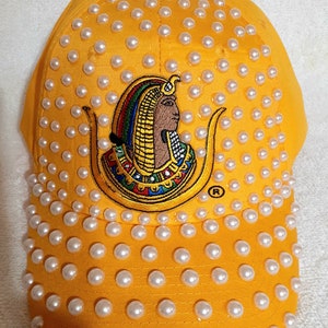 Könnte beinhalten: Eine gelbe Baseballkappe mit weißen Perlenbesatz. Die Kappe zeigt ein gesticktes Bild eines ägyptischen Pharaos mit einem bunten Kopfschmuck und dem Buchstaben "R" in einem Kreis.