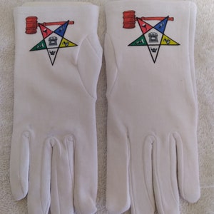 Puede incluir: Un par de guantes de algodón blanco con un símbolo masónico bordado en la parte posterior de cada guante. El símbolo es una estrella de cinco puntas con una brújula y una escuadra en el centro.