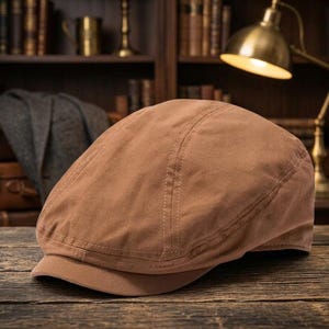 May include: A solid tan flat cap constructed from a smooth, matte fabric. The cap features a rounded crown with visible stitched panel seams and a short, stiffened brim extending from the front, maintaining a structured, low-profile shape.
