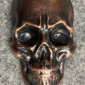 May include: A three-dimensional skull sculpture featuring a dark, weathered bronze finish with highlighted copper-toned edges along the brow, cheekbones, and jawline, showcasing a smooth, polished surface texture throughout the entire piece.