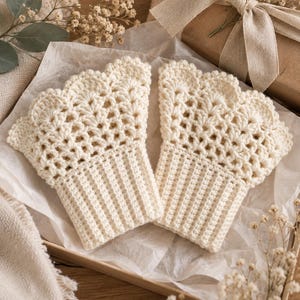 Könnte beinhalten: Ein Paar fingerlose Handschuhe in Off-White, gefertigt in Häkeltechnik. Die Handschuhe verfügen über ein geripptes Bündchen am Handgelenk und ein komplexes, durchbrochenes Muschelmuster, das sich zu den Fingern hin erstreckt und einen gewellten Rand bildet.