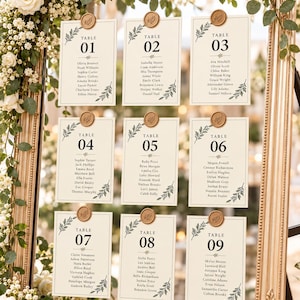 Puede incluir: Un plano de mesa de boda con nueve tarjetas de color crema dispuestas en cuadrícula sobre un espejo con marco dorado. Cada tarjeta muestra un número de mesa del 01 al 09, nombres de invitados y una ilustración botánica, fijada con un sello de cera dorado.