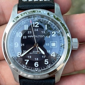 May include: A Hamilton Khaki Automatic wristwatch with a black dial, silver-tone hands, and white Arabic numerals. The watch features a date window at the three o'clock position, a black leather strap with white stitching, and a stainless steel case.