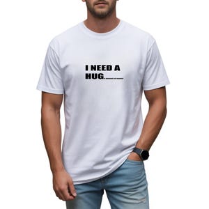 May include: A white short-sleeved t-shirt featuring the black printed text I NEED A HUG...e amount of money centered on the chest. The garment has a crew neckline and a standard fit, providing a clean and simple look for casual daily wear.
