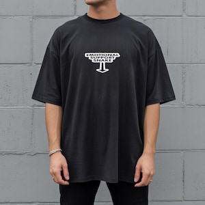 May include: A black short-sleeved t-shirt featuring a white graphic on the chest. The graphic displays the text "EMOTIONAL SUPPORT SNAKE" arranged in a block shape with a downward-pointing arrow underneath the lettering, centered on the front.