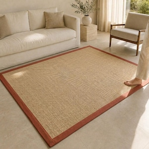 May include: A rectangular area rug featuring a natural tan woven fiber center with a solid terracotta-colored fabric border. The rug lies flat on a light-colored floor, showcasing a textured, tight-knit surface pattern throughout the main area.