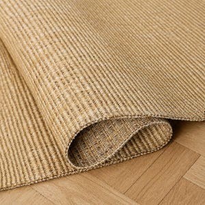 May include: A tan-colored area rug featuring a dense, woven texture with a consistent ribbed pattern. The material displays a natural, earthy tone with subtle variations in light and dark beige fibers, creating a uniform and structured appearance.