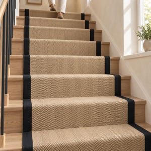 May include: A stair runner featuring a tan herringbone pattern with solid black fabric borders on both sides. The runner is installed on wooden stairs, covering the center of each tread and riser, creating a continuous path up the staircase.