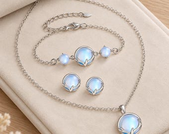 Könnte beinhalten: Ein Schmuckset bestehend aus einer silberfarbenen Halskette, einem Armband und einem Paar Ohrsteckern. Jedes Stück zeigt einen runden, schimmernden blauen mondsteinartigen Cabochon in einer silbernen Fassung mit Metallverzierungen.