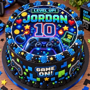 Könnte beinhalten: Ein runder Kuchen mit schwarzer Glasur, verziert mit blauen und schwarzen Cremetupfern und bunten Sternstreuseln. Die Oberseite zeigt eine Videospielgrafik mit dem Text "LEVEL UP!", "JORDAN", "10" und "GAME ON!" sowie einen Gamecontroller.