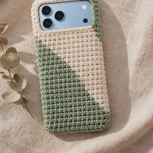 Può includere: Una custodia per telefono rettangolare caratterizzata da un design all'uncinetto bicolore in verde salvia e crema. La custodia è realizzata con un motivo in filato intrecciato e testurizzato che copre il retro e i lati, lasciando un'apertura per le lenti.
