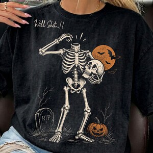 Peut inclure: Un t-shirt noir avec un motif de squelette sans tête tenant son propre crâne. Le dessin comprend une pierre tombale avec le texte RIP, une citrouille et une lune orange avec des chauves-souris. Le texte Will Shit..!! est écrit en blanc.