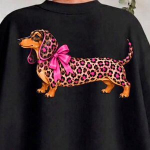 Peut inclure: Un sweat-shirt noir orné d'un motif de teckel au visage et au corps fauves, recouvert d'un imprimé léopard rose et noir. Le chien porte un grand nœud rose vif autour du cou, centré sur le dos du vêtement.