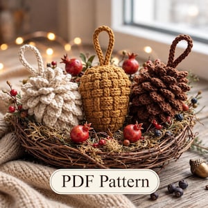 Könnte beinhalten: Drei gehäkelte Ornamente in Form von Tannenzapfen in Creme, Senfgelb und Dunkelbraun, jeweils mit einer Aufhängeschlaufe. Die Ornamente sind in einem geflochtenen Nest mit kleinen roten Früchten und grünem Moos arrangiert. Text: PDF Pattern.