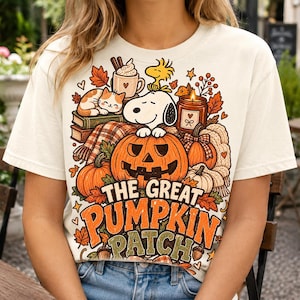 May include: A cream-colored t-shirt featuring a graphic print of Snoopy and Woodstock sitting on a jack-o'-lantern surrounded by autumn elements including pumpkins, a cat, a mug, a candle, and leaves with the text The Great Pumpkin Patch.
