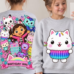May include: A light gray sweatshirt featuring a colorful graphic of a girl with cat ears surrounded by various cartoon cats, a guitar, and a building, with a pink banner reading BIRTHDAY GIRL. A second sweatshirt shows a white cat cupcake graphic.