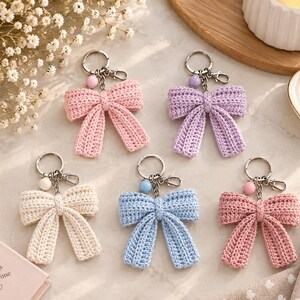 Può includere: Cinque portachiavi a forma di fiocco all'uncinetto in colori pastello, tra cui rosa chiaro, lavanda, crema, azzurro e rosa antico. Ogni fiocco presenta un anello portachiavi in metallo argentato, un piccolo moschettone e una perlina.