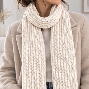 May include: A thick, cream-coloured ribbed knit scarf worn around the neck, featuring a vertical textured pattern. The scarf is draped over a beige coat and a white top, with the ends hanging down straight in front of the torso.