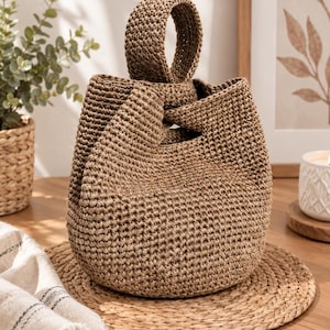 Peut inclure: Un sac seau marron crocheté à la main avec une base arrondie et une poignée circulaire unique intégrée. Le sac présente un motif de points dense et texturé sur toute sa structure, créant une apparence uniforme et solide avec une ouverture.