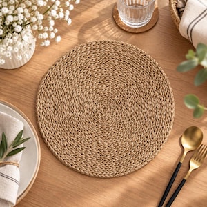 Puede incluir: Un mantel individual redondo trenzado hecho de fibras naturales de color tostado con una textura apretada. El mantel presenta un patrón circular que irradia desde el centro, creando una superficie consistente y uniforme.