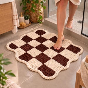 May include: A square-shaped bathroom rug featuring a dark brown and cream checkerboard pattern. The rug has a scalloped, wavy edge finished with a thick cream-coloured border. The texture appears to be crocheted with a chunky yarn construction.