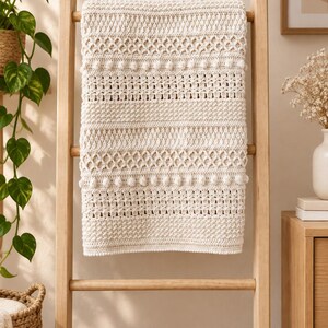 May include: A cream-colored crocheted blanket featuring a repeating pattern of textured bobbles, diamond-shaped openwork, and horizontal bands of dense stitching. The rectangular textile is draped over a light-toned wooden ladder frame.