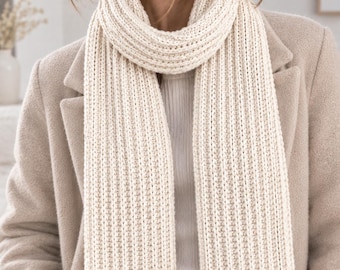 Peut inclure: Une écharpe en tricot côtelé de couleur crème portée autour du cou, présentant un motif vertical épais et texturé. L'écharpe est faite d'un fil épais et doux, et tombe en deux longs pans droits sur un manteau beige et un haut blanc côtelé.
