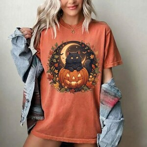 May include: A burnt orange t-shirt featuring a graphic of a black cat sitting on a carved jack-o'-lantern pumpkin, surrounded by a circular frame of autumn leaves and a crescent moon, worn with a light blue denim jacket.