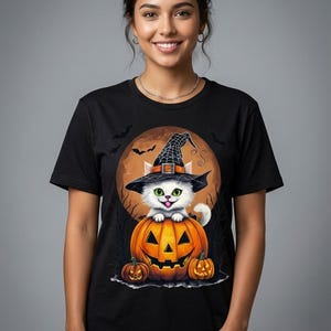 May include: A black t-shirt featuring a graphic print of a white kitten wearing a black witch hat, sitting atop a large orange jack-o'-lantern. The design includes two smaller jack-o'-lanterns, a full moon background, and silhouettes of flying bats.