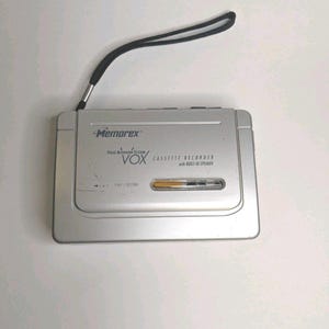 May include: A silver Memorex cassette recorder featuring a voice activated system, a built-in speaker, and a black wrist strap. The front panel displays the text Memorex, Voice Activated System VOX, Cassette Recorder with built-in speaker, and Play/Record.