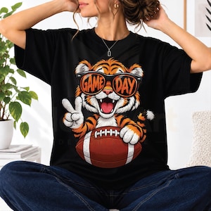 Puede incluir: Una camiseta negra con un gráfico de un tigre que lleva gafas de sol con el texto GAME DAY impreso en los cristales. El tigre sostiene un balón de fútbol americano marrón y hace el signo de la paz con una pata. El diseño está centrado.
