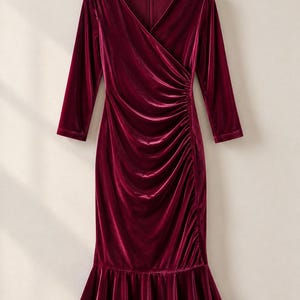 May include: A deep burgundy velvet dress featuring three-quarter length sleeves, a V-neckline, and a wrap-style bodice with side ruching. The garment has a fitted silhouette that transitions into a flared, ruffled hemline at the bottom.