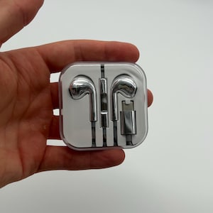 May include: A pair of shiny silver wired earbuds with a matching silver connector plug, neatly coiled and secured inside a clear, square-shaped plastic protective carrying case with rounded corners and a visible internal white plastic organizer.